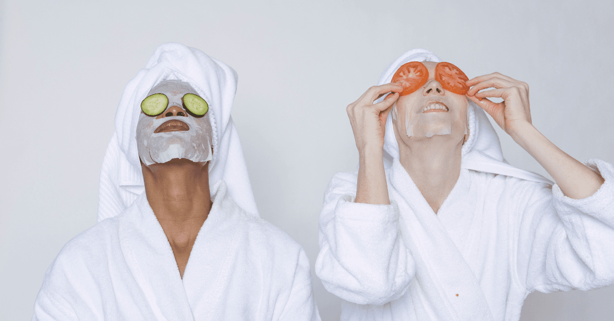 Zwei Personen mit Gesichtsmasken und Handtüchern auf dem Kopf entspannen mit Gurken- bzw. Tomatenscheiben auf den Augen.